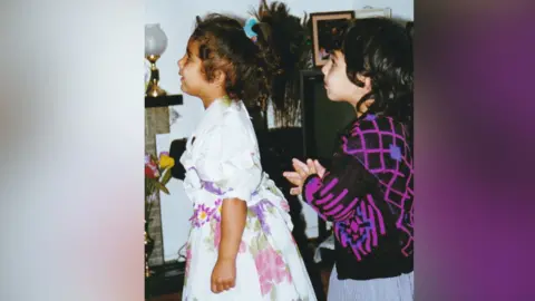 Jussi Sidhu Childhood photo of Jussi and her sister. you girl British Indian girl on the right wearing a white and pink and purple flora dress with puffy sleeves. Her hair curly hair is tight up worth a blue srunchy. The girl on the right is British Indian and with short dark hair and a fringe, wearing a black and purple zig-zag patterned cardigan.