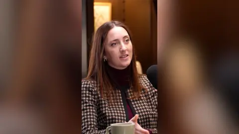 A woman with long, brown hair talks to someone off-camera