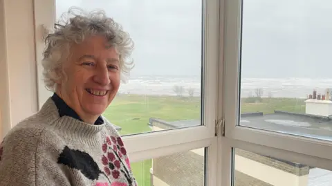 Helen Coyne is in her seventies and is wearing a beige jumper with a pattern of pink and purple flowers on it. She is standing by a window with white uPVC frames, through which some grass and the sea beyond can be seen. The day looks a bit grey.