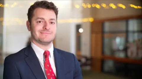 Jamie Niblock/BBC Labour MP for Ipswich Jack Abbott smiles at the camera inside a building. He has short dark hair and wears a navy suit, white shirt and red tie. 