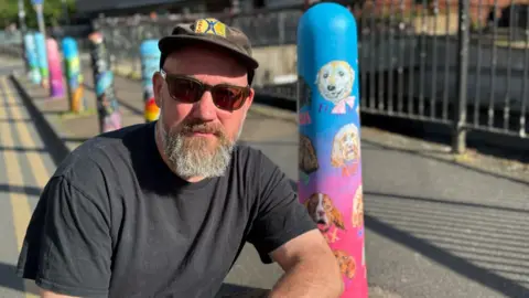 A man crouching next to a bollard painted blue and pink with portraits of dogs.