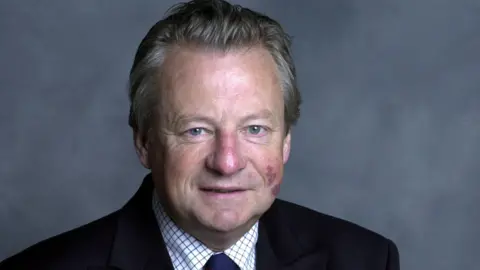 BBC Dafydd Elis-Thomas who has grey hair and a birthmark on his cheek. He is wearing a black suit and is smiling and looking at the camera.