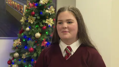 A girl in a red V-neck jumper, white shirt and red, green and grey striped tie. Their is a green Christmas tree decorated with red and gold stars as well as red, blue and green lights. The wall is a light-yellow colour. 