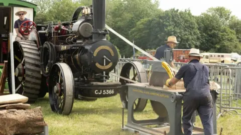 Gorman Photography The photo shows a black locomotive being driven by a man in a wide-brimmed hat and sunglasses. In front of it another man cuts a piece of wood with a table saw. 
