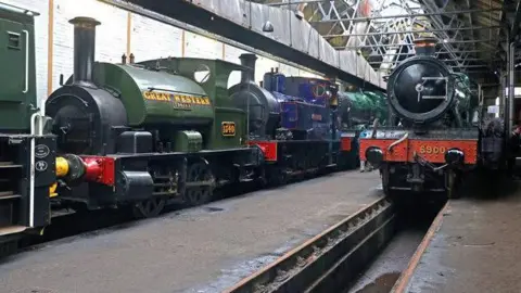 Four vintage steam engines in an engineering shed. One on the left is racing green with the words 'Great Western' in yellow on the side. Another black engine has the number 5900 on a red panel on its front.