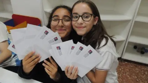 Ines and Sabela look at the camera holding up several letters. Both girls have glasses and brown hair.