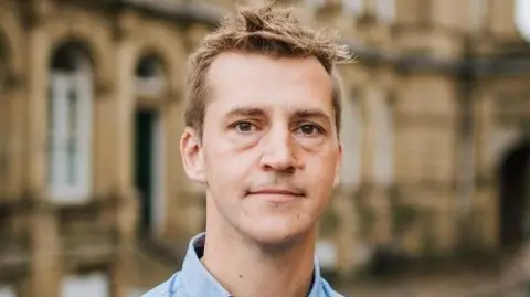 LDRS Calderdale's Labour MP Josh Fenton-Glynn - a white man with blonde hair and a blue shirt standing in front of sandstone buildings