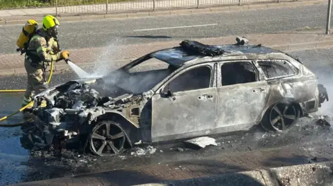 Scott Chisholm Firefighter in protective gear hosing down a blackened car which is burnt-out on a road