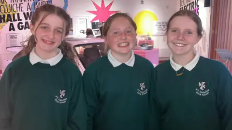 Zoe, Bella-May and Kate at the 80s exhibition. They are wearing green school jumpers over white shirts. Zoe has long brown hair while the others have shorter brown hair pulled back behind their heads.