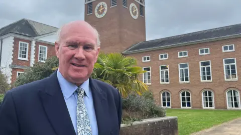 James McInnes, leader of Devon County Council, wearing a blue shirt and patterned tie and a blue blazer.