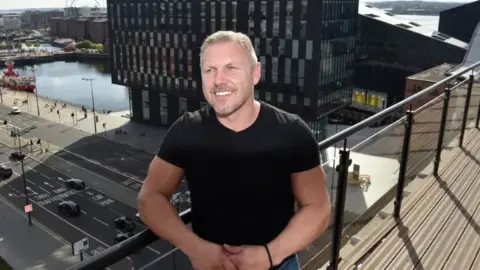 A man in his 50s, with short blonde hair and light stubble, poses on the balcony of tall building overlooking an area of dockland. He is wearing a black t-shirt and is smiling. 