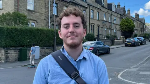 LDRS A younger man with curly brown hair and a blue shirt stands on a residential street.