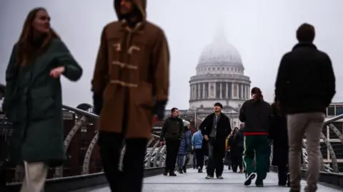 Getty Images Pedestrians walk across the Millennium Bridge on a cloudy day, with the dome of St. Paul's Cathedral partially hidden in the mist in the background. People in various winter outfits.