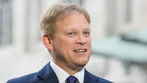 Grant Shapps has fair hair and is wearing a navy jacket over a white shirt and blue and pink tie. He is smiling with his mouth open.
