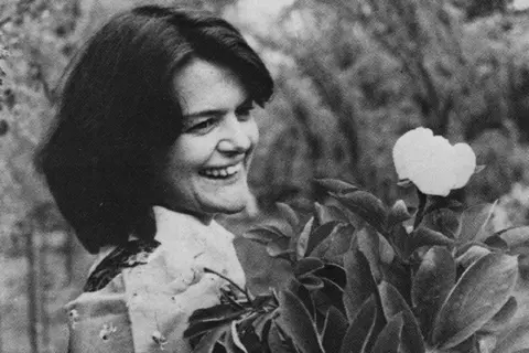 Maggie's Centres Black-and-white image of Maggie Keswick Jencks. She has dark hair and is smiling at something off camera. In front of her is a flowering plant. 