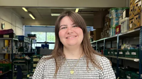 Kate McIntosh is smiling at the camera while in the food bank. She is wearing a blue and white checkered top and a gold necklace. She has long brown hair and a side fringe. Behind her are shelves with green trays on. In the centre of the room, also behind her, are more trays stacked up with food inside them.