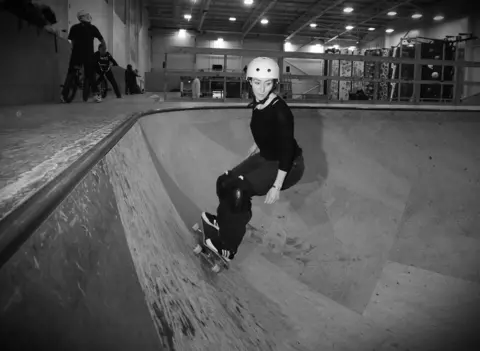 Nathan Livingston Black and white image shows a girl in black on skateboard with white helmet on wooden ramp