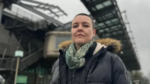 Campaigner Jo Howell stands in front of the National Glass Centre on the banks of the River Wear, wearing a thick waterproof jacket and a scarf. She has short, dark hair.
