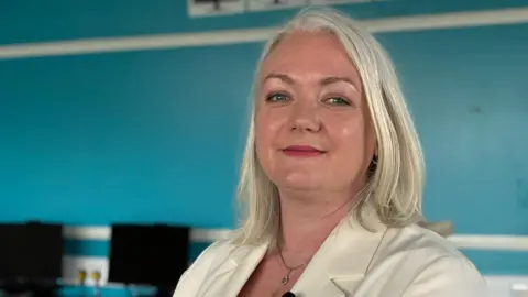 A woman with white grey shoulder-length hair. She is wearing a white blazer. She is stood side-on and looking at the camera with a small smile.