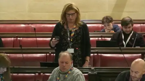 Bristol City Council/YouTube Kaz Self standing at a council meeting. She has blonde hair and is wearing a black dress and a black cardigan. Several other councillors are sitting on the benches.