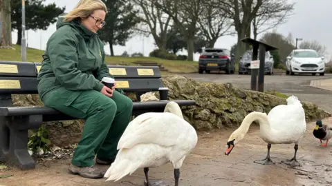 Michaela Jackson A woman with blonde hair wears glasses and a green uniform as she sits on a dark wooden bench in a park. She is holding a disposable coffee cup and looking at two white swans and a mallard duck, which are standing on the pavement in front of her. There is a small rock wall and three cars parked in the distance.