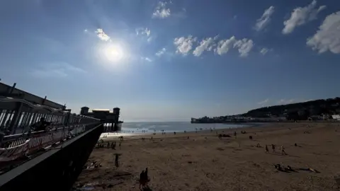 The dark outline of Weston Pier is on the left of the image. A beach and the sea beyond feature several dozen people in the distance, with a headland on the right.