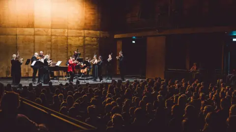 A group of musicians on stage, most are wearing black but a musician in the centre is wearing a red jacket and playing the violin. There are more musicians playing violins, with a piano and viola as well. The stage wall behind them is wooden and lit up in a yellow light. The audience are sitting down in rows and the backs of their heads are in shadow. It looks very full.