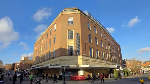 The exterior of the Debenhams building in Norwich on a sunny day. It is a multi-storey building, constructed in the 1950s. Shoppers are walking past it.
