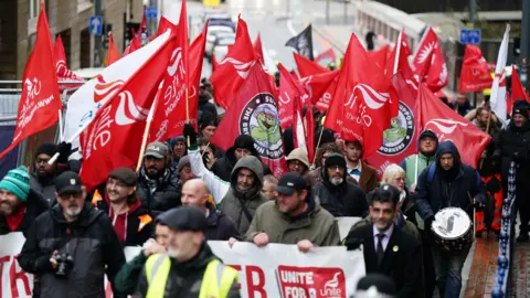 Striking workers marching along a Birmingham street. Some are holding a banner, others carrying red and white Unite the union flags
