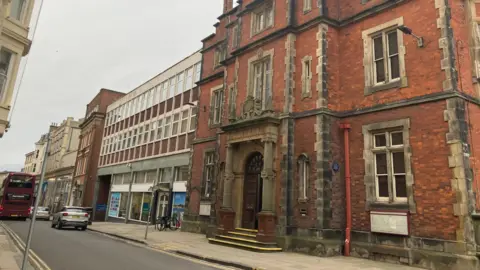 The image shows a street scene in an urban area, featuring a mix of historic and mid‑20th‑century buildings. The weather appears overcast, giving the scene muted lighting. A narrow asphalt road runs through the scene, with double yellow lines along the left edge, indicating no parking. A pavement runs alongside the buildings, made of light grey stone slabs. A large, ornate historic building dominates the right side of the image.