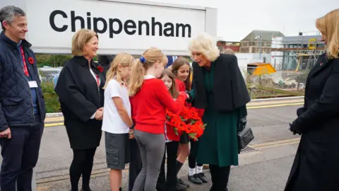 PA Media Primary school pupils hand Queen Camilla a poppy wreath made from crochet. She is speaking to one of them and a few adults dressed in black surround them. They are on a Chippenham train platform, with the big sign for the town behind them.