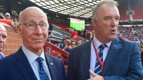 John Shiels Sir Bobby Charlton is standing on the left, wearing a navy blue suit and tie with a pale blue shirt. He is largely bald and wears glasses. John is on the right also wearing a blue navy suit. Crowds in the stadium are in the background.  