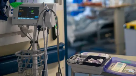PA Media Hospital equipment with a monitoring screen and a bed with what appears to be needles and further monitoring equipment on it. A hospital room, including a bed, can be seen in the blurred background. 