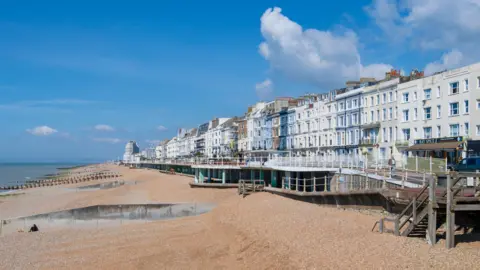 A view of Hastings seafront, featuring its shingle beach, promenade and Victorian terrace buildings.