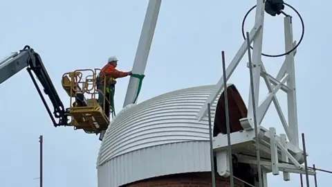 Adam Singer A man in a cherry picker works on one of the stocks of the Toft Monks Wind Pump, which features a white weather boarded cap, with tail fan structure to the right of the image.