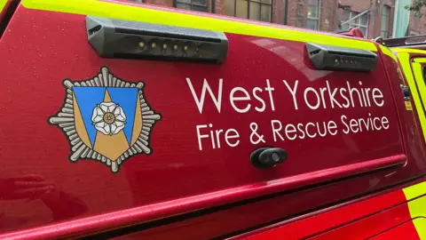 The side of a bright red emergency vehicle belonging to the West Yorkshire Fire & Rescue Service. The text “West Yorkshire Fire & Rescue Service” is clearly printed in white letters on the vehicle’s surface. To the left of the text, there is an official emblem featuring a white rose set against a blue and gold shield, surrounded by a stylized silver starburst design.
The vehicle has high-visibility markings, including fluorescent yellow stripes along the bottom edge. Two black rectangular light units with multiple LED bulbs are mounted on the top section of the vehicle. In the background, there are brick buildings with large windows, some of which are illuminated.