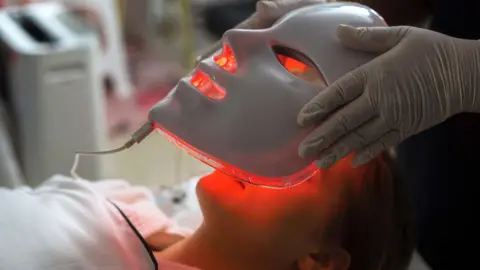 A white red light therapy face mask is being carefully placed on a woman's face she lies down