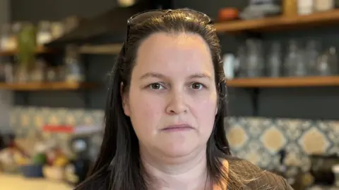 A woman stands in a kitchen. She has long brown hair and is wearing a brown top