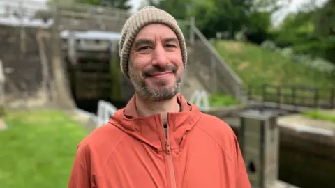 Olivia Richwald/BBC A man stands in front of a canal lock. He is wearing an orange coat and hat as he smiles at the camera.