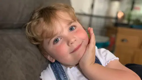 The Milnes family Amber is smiling at the camera as she leans her head back on a sofa and rests her chin on her left hand. She has clear blue eyes and long blonde hair tied back from her face. She is wearing a short-sleeved white T-shirt and there are two denim straps over a shoulder. 