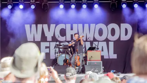 Wychwood Festival A violinist plays on a stage with Wychwood Festival written behind him and a crowd watching on a bright sunny day