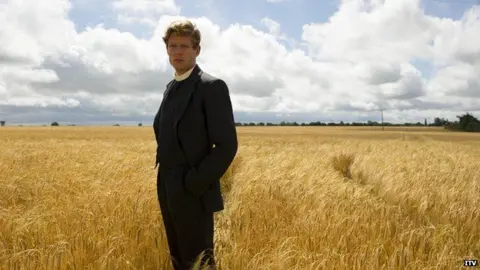 ITV James Norton dressed as an Anglican clergyman in the 1950s and standing in a golden corn field. He is wearing a black suit, with a white dog collar and looking towards the viewer. His hands are in his pockets. Beyond the flat field, it is edged with trees and above is a cloud covered blue sky.