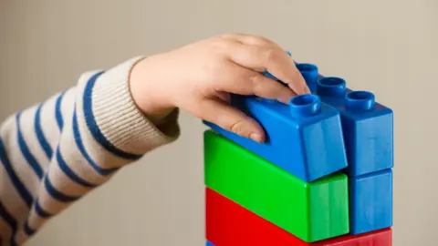 A stock image of a child's hand stacking large colourful Lego blocks. Only the arm and hand of the child are visible. They are wearing a striped cream and blue jumper. There are red, green and blue Lego blocks stacked to make a tower.