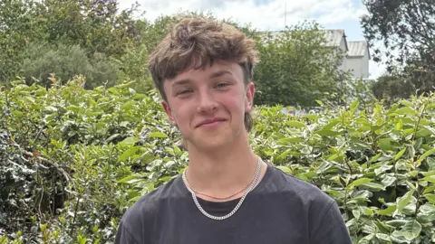 Ethan has long wavy brown hair and wears a black t-shirt with two chains around his neck. 