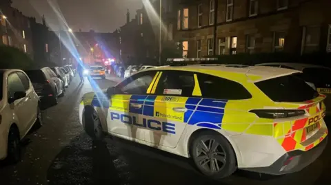 A blue and yellow marked police care parked across a street in Glasgow. Another car can be seen parked in the distance. The street is lined with tenement buildings.