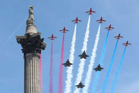 Getty Images The Red Arrow take part in the flypast over Buckingham Palace. Twelve aircraft are pictured with plumes of red, white and blue clouds in their wake through a blue sky.