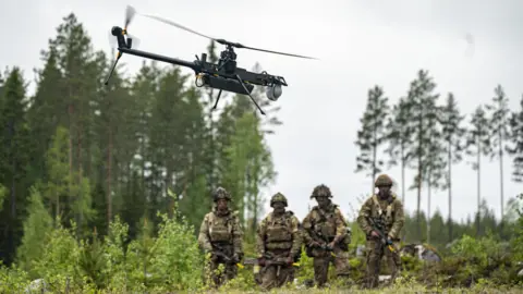 Four British Army soldiers stand in a wooded area watching a single-rotor helicopter style Ghost drone