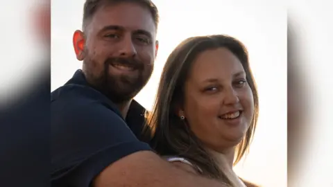 Becca Moore and her male partner are stood together outside. She is stood with her back to him and he has his arms over her shoulders. They are both smiling at the camera. Her partner has a blue t-shirt and brown hair and facial hair. Becca has long brown hair with blonde highlights and wears earrings.   