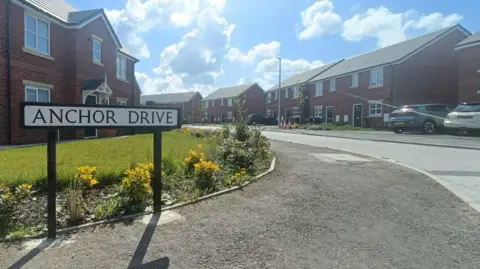 A sign reads Anchor Drive as the road leads round into a residential area - with houses on both sides of the road and cars in the drive