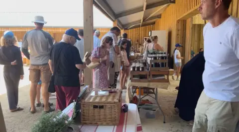 Visitors browse stalls inside a covered market space with wooden walls and support beams. Tables display jars, bottles and local produce while shoppers queue and move between vendors. Natural light fills the open-sided building, creating a busy community market atmosphere.